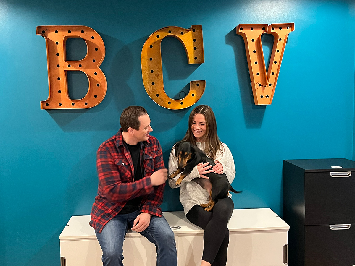 Two BCV team members sitting on a bench on the office holding a puppy with BVC on the wall behind them in light up letters