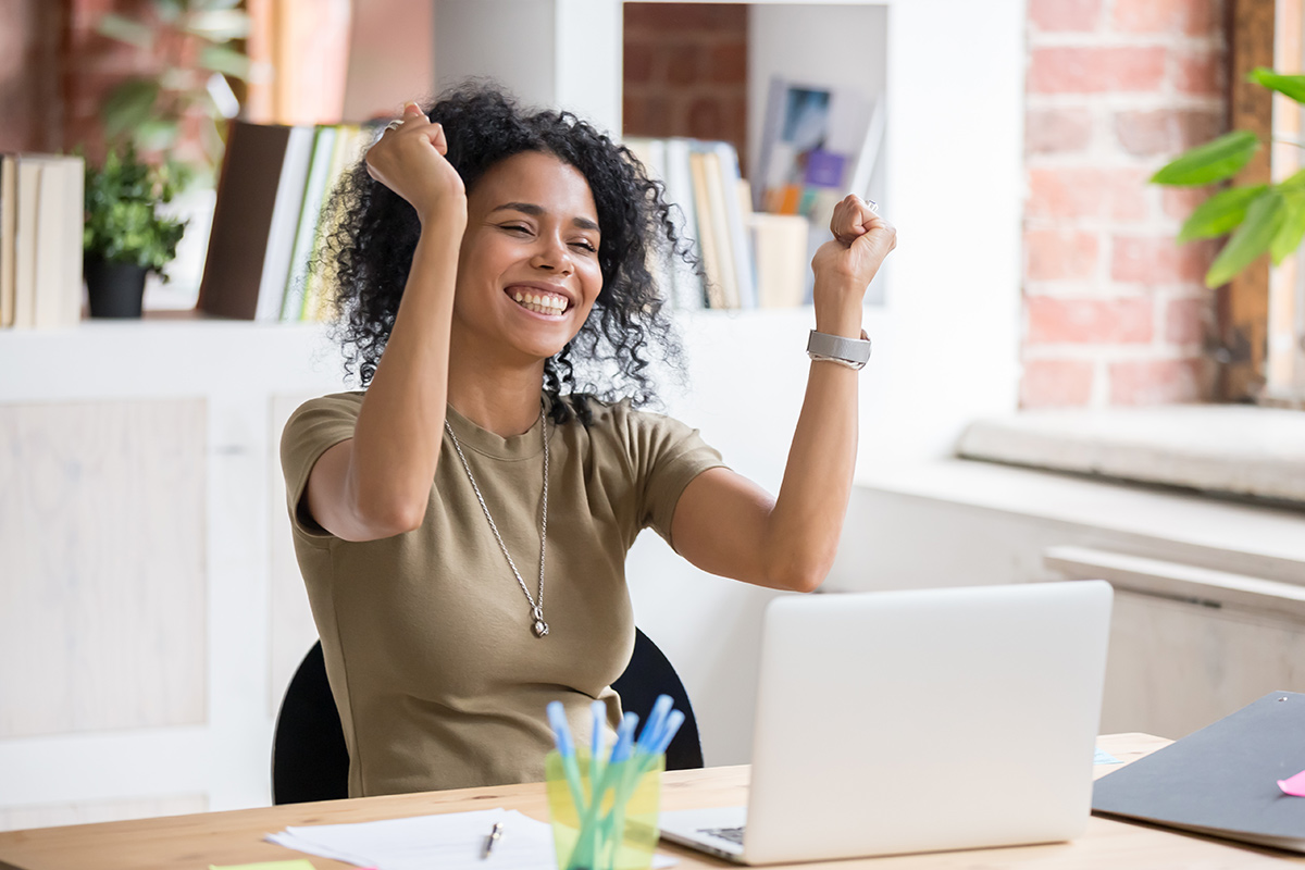 excited biracial girl sitting at a desk looking at a laptop with her arms raised in celebration