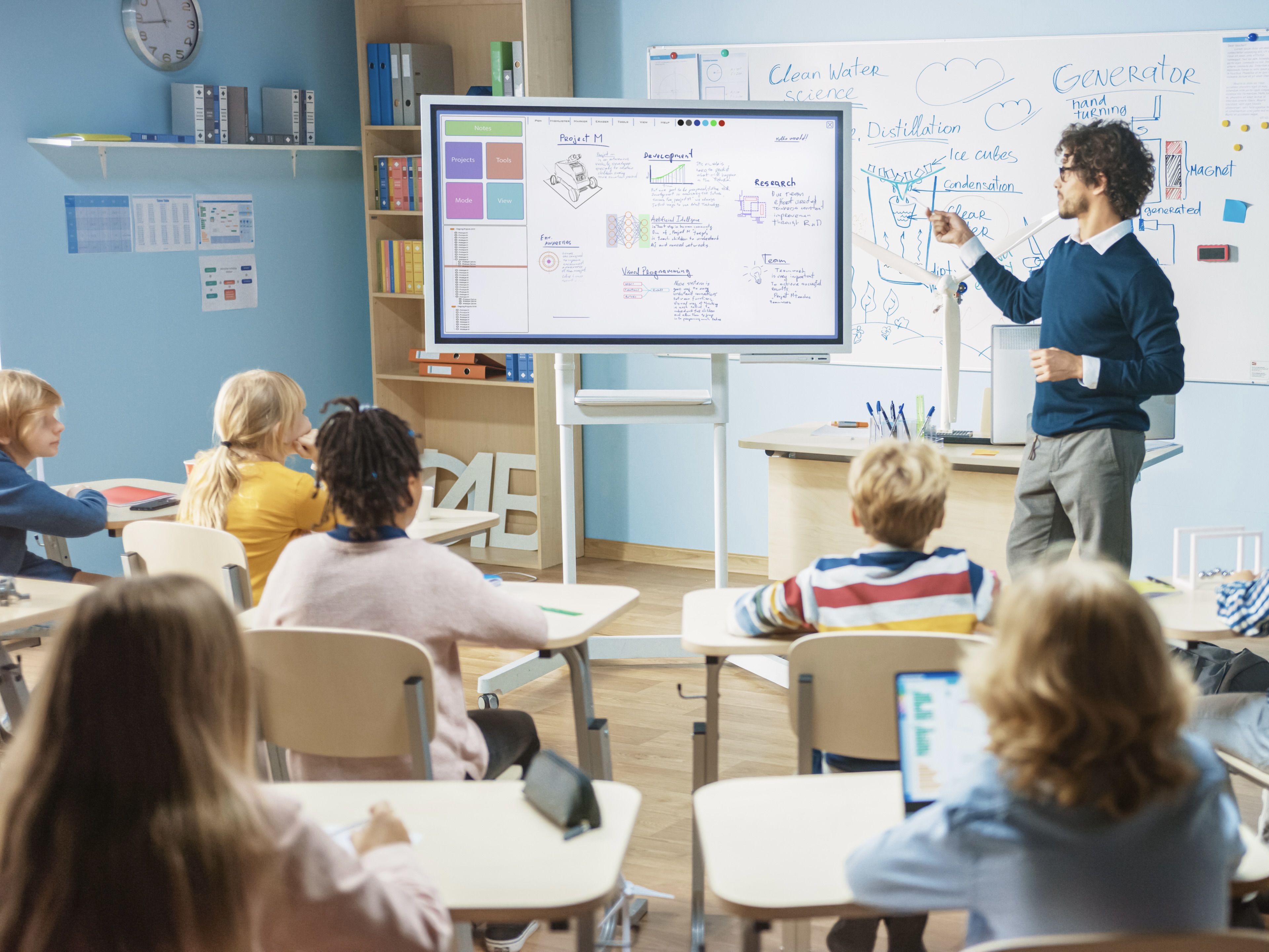 A classroom of children looks at a digital display as their teacher indicates something on the board.