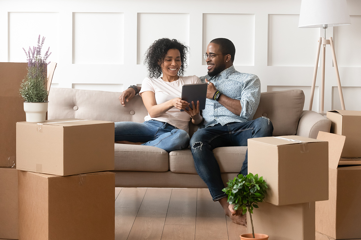 Happy couple sitting on a couch looking at a digital tablet with moving boxes on the floor