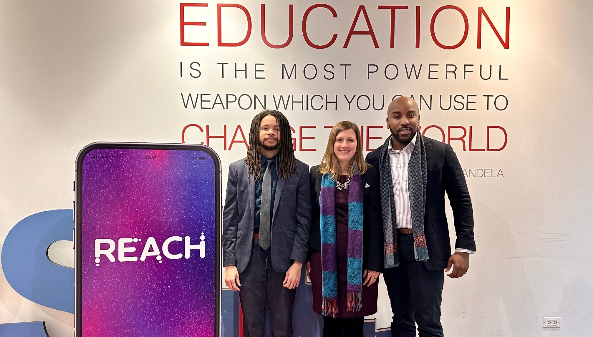 The Reach Pathways founding team poses together for a photo next to a large mobile phone displaying the REACH app