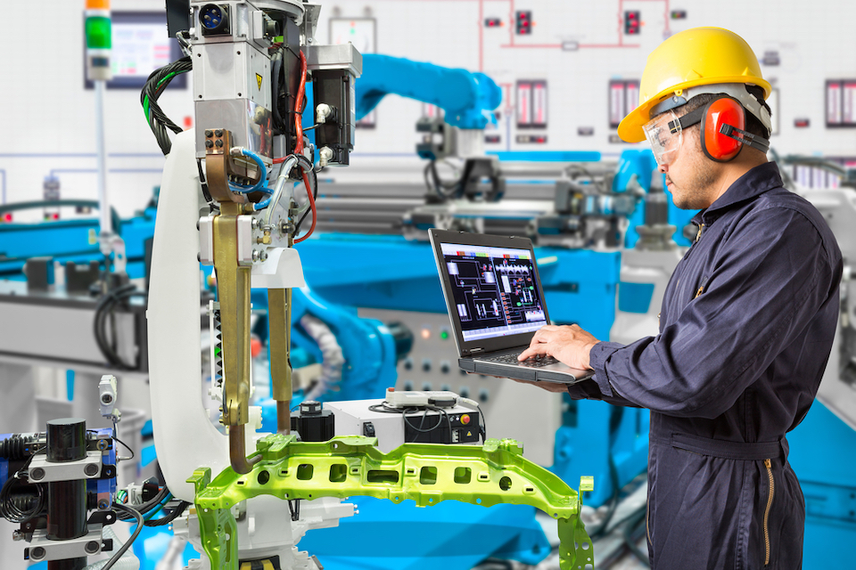 Engineer checks laptop while robotic arms manufacture a product