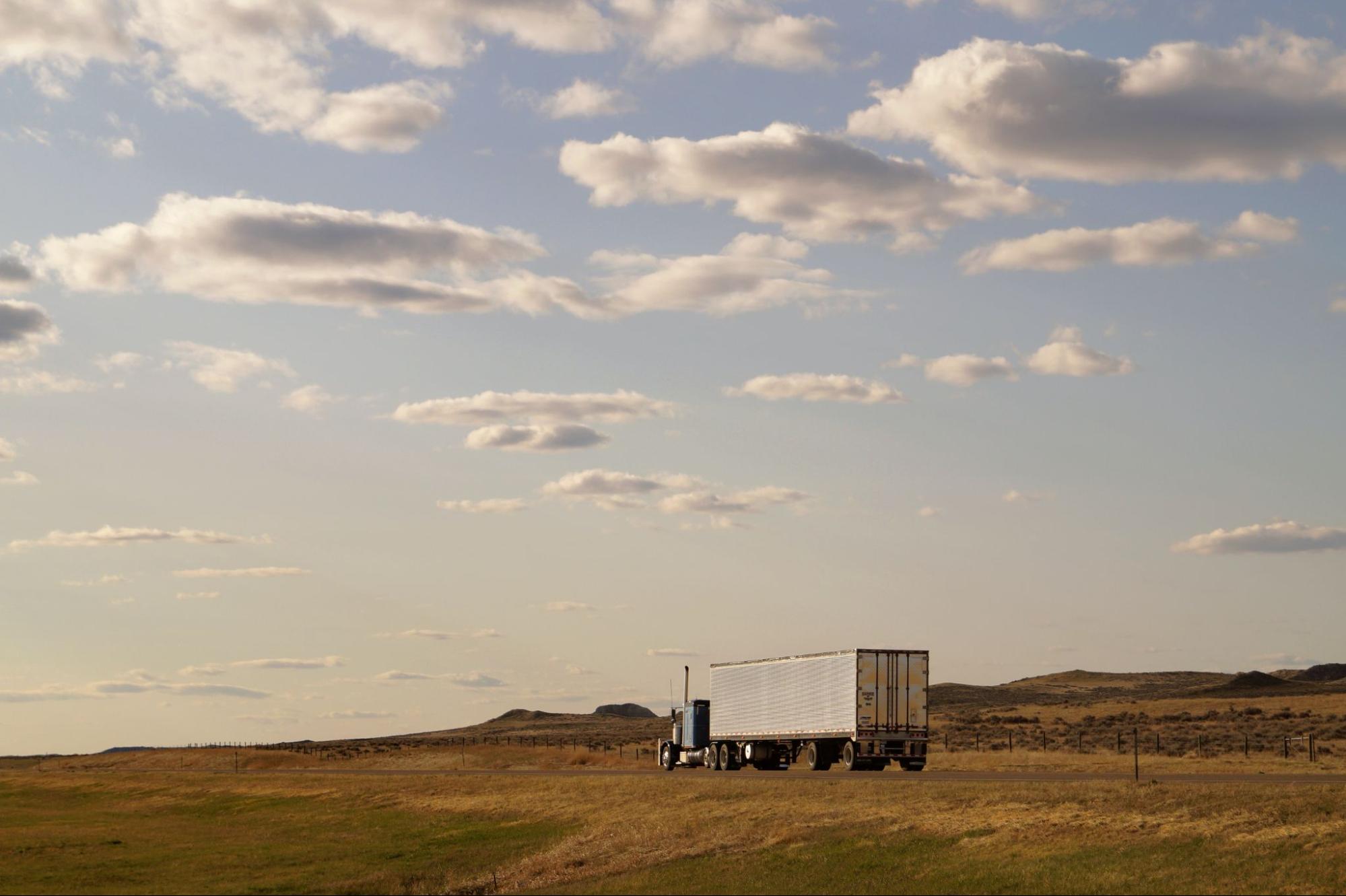 A semi truck driving along a road surrounded by open fields and clear sky.