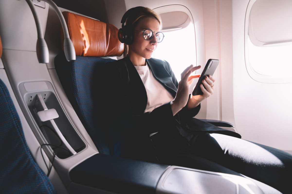  A woman in a business suit with headphones on uses her smartphone on an airplane.