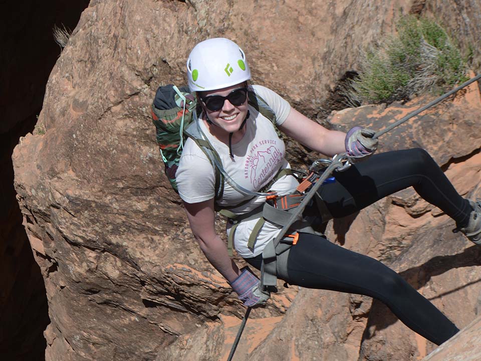 Associate Manager of Research Services Olivia Benson rock climbing, looking up the cliff at the camera and smiling.