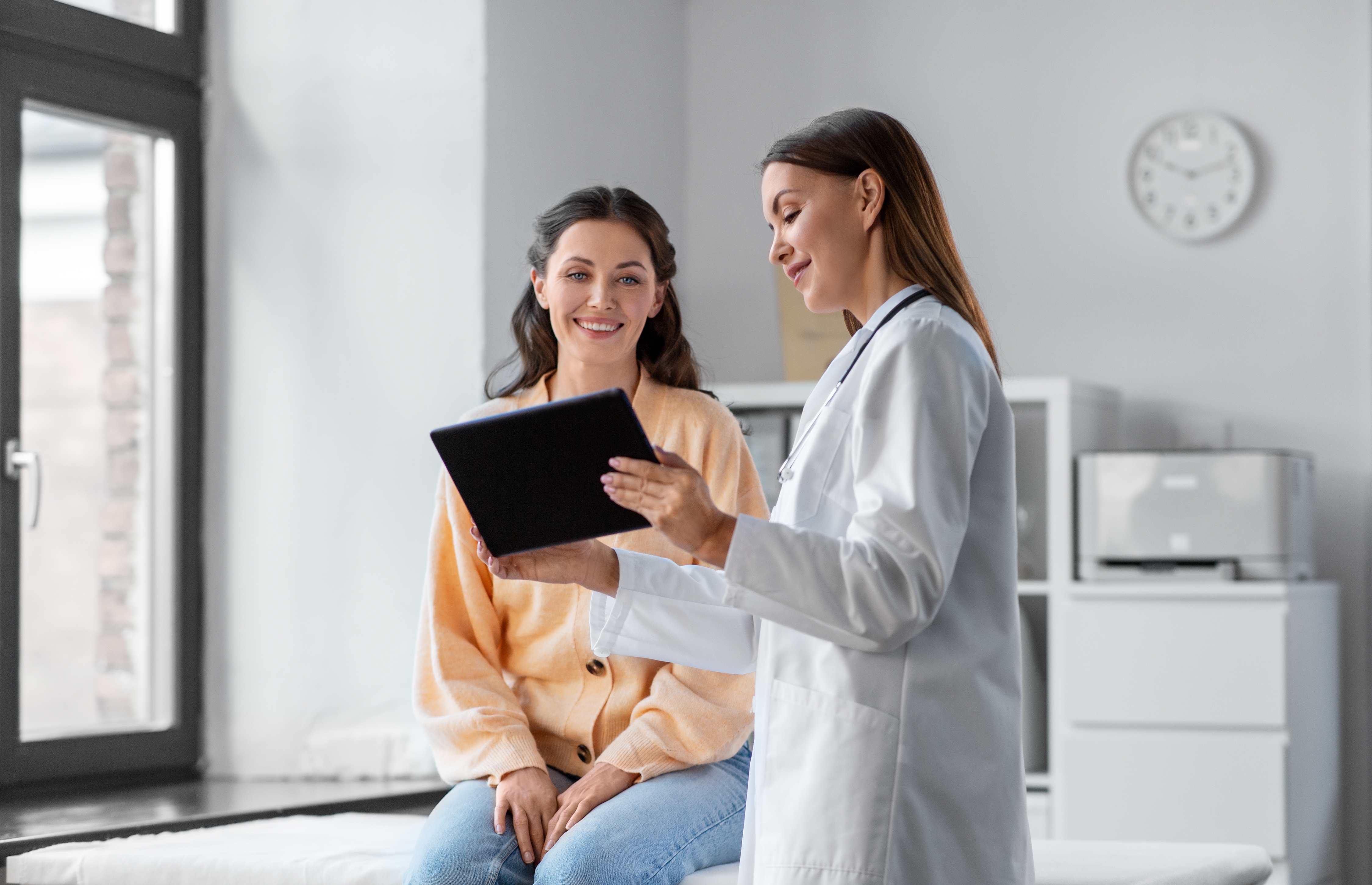 A young woman meets with their doctor in a clinical setting; the two review information on a tablet