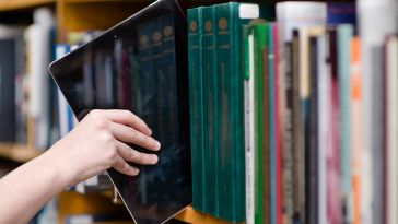 A student pulling a tablet from a bookshelf.