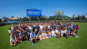 Large group photo of GoHealth team in Wrigley Field’s outfield with GoHealth logo on billboard behind them